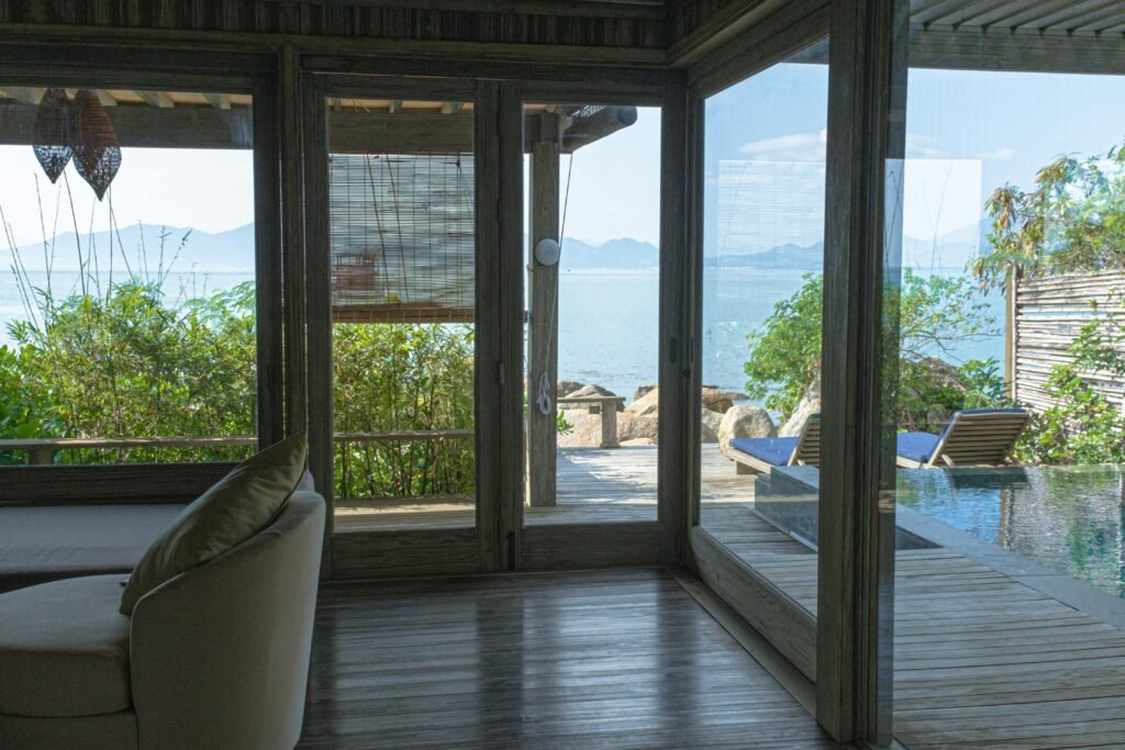 Stunning beachfront view from a wooden framed room with glass doors, overlooking the ocean and lush greenery.
