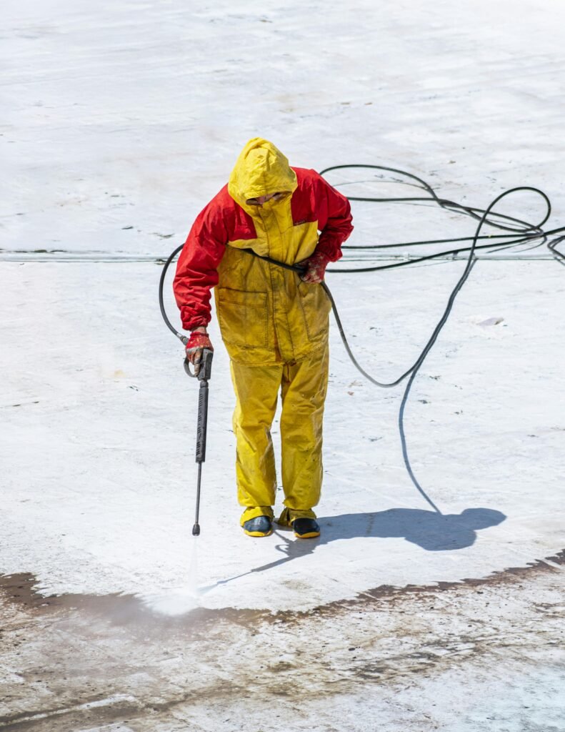 Person in yellow and red safety suit cleaning with a pressure washer outdoors.